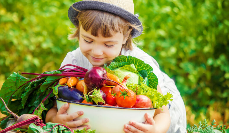 O fetiță cu pălărie ține în brațe un bol în care se află legume.