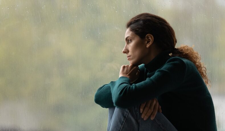 Femeie tristă care privește pe geam, cu stare depresivă