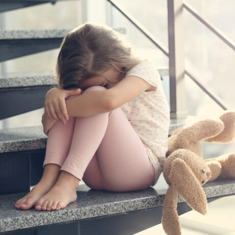 O fetiță care stă cu o jucărie alături, pe niște scări din beton, cu capul pe genunchi