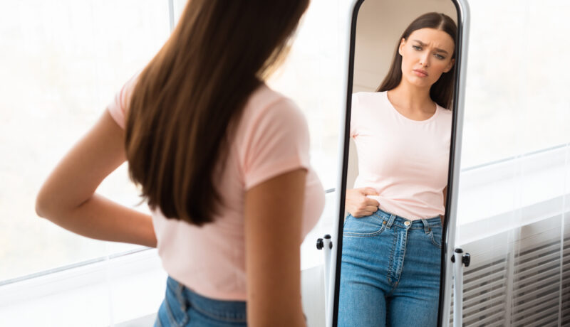 O femeie care se uită în oglindă și își vede grăsimea de pe abdomen