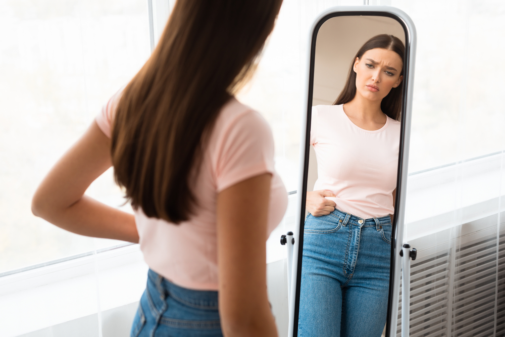 O femeie care se uită în oglindă și își vede grăsimea de pe abdomen