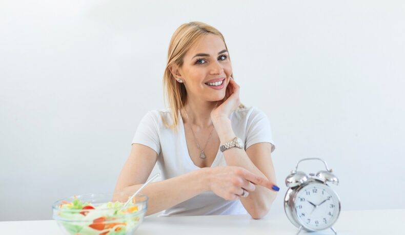 Femeie la masă care arată ceasul și are o salată pe masă, cu referire la postul intermitent