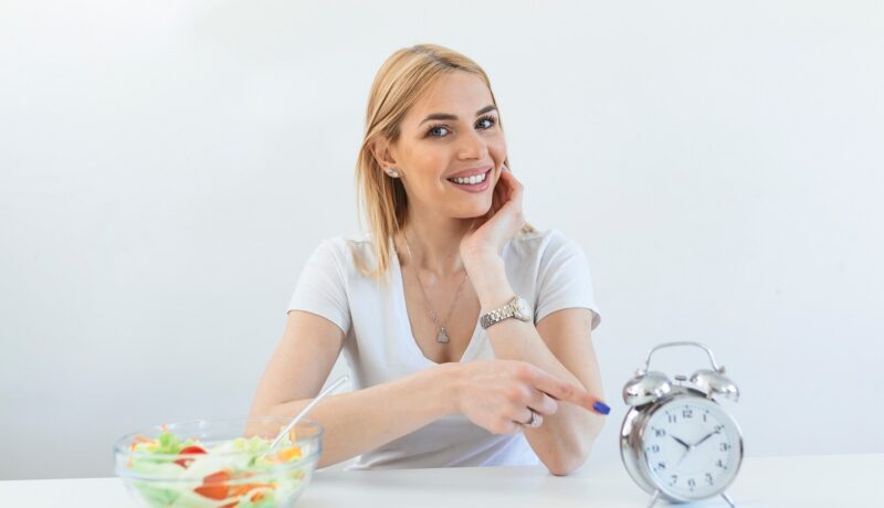 Femeie la masă care arată ceasul și are o salată pe masă, cu referire la postul intermitent