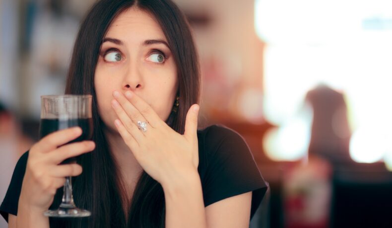 Femeie cu mâna la gură care sughiță și ține în mână un pahar cu suc de culoare închisă