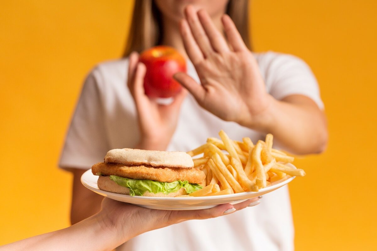 Femeie cu un măr în mână care refuză o farfurie cu cartofi și hamburgeri, alimente care produc inflamație în corp