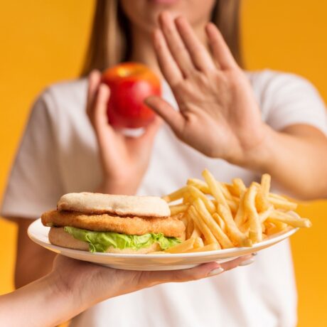 Femeie cu un măr în mână care refuză o farfurie cu cartofi și hamburgeri, alimente care produc inflamație în corp