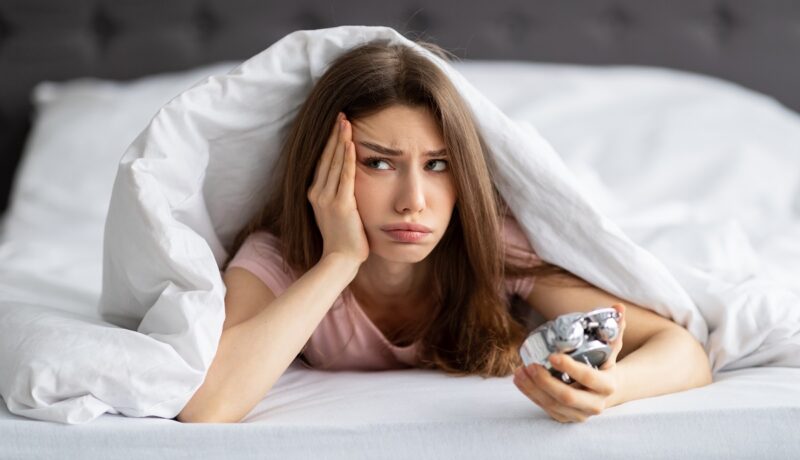 Fata care stă în pat, se uită la ceas și nu poate să adoarmă, în căutare de soluții pentru cum să adormi mai repede