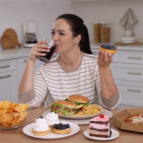 Femeie care bea cola și mănâncă o tartă, cu burgeri, cartofi și părjituri pe masă - toate fiind alimente care scad imunitatea