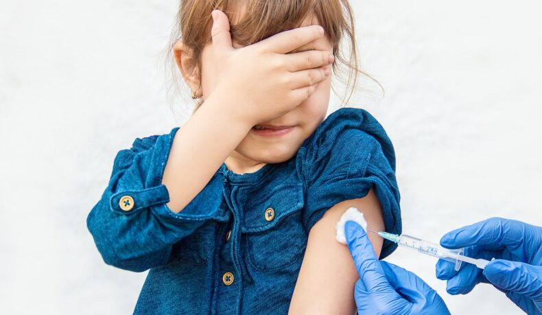 Fetiță care primește un vaccin în braț și își acoperă ochii cu mâna, sugestiv pentru vaccinul antivaricelă la copii
