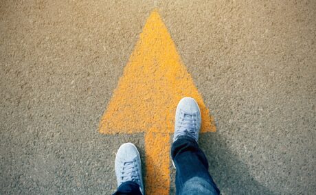 Picioare care stau pe o săgeată galbenă desenată pe asfalt, sugestiv pentru mersul pe jos