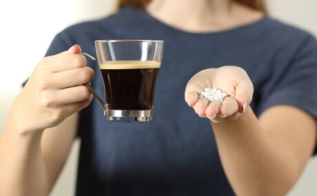 Femeie care ține în mână o cafea și tablete de îndulcitori artificiali, două lucruri care pot duce la un exces de zahăr în sânge