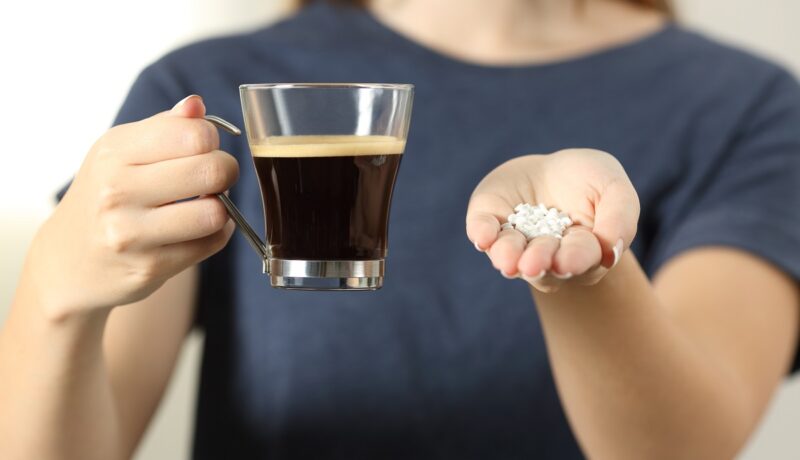 Femeie care ține în mână o cafea și tablete de îndulcitori artificiali, două lucruri care pot duce la un exces de zahăr în sânge
