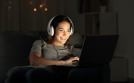 O femeie tânără, cu o pereche de căști pe cap, zâmbește și se uită la ecranul unui laptop noaptea, într-o cameră
