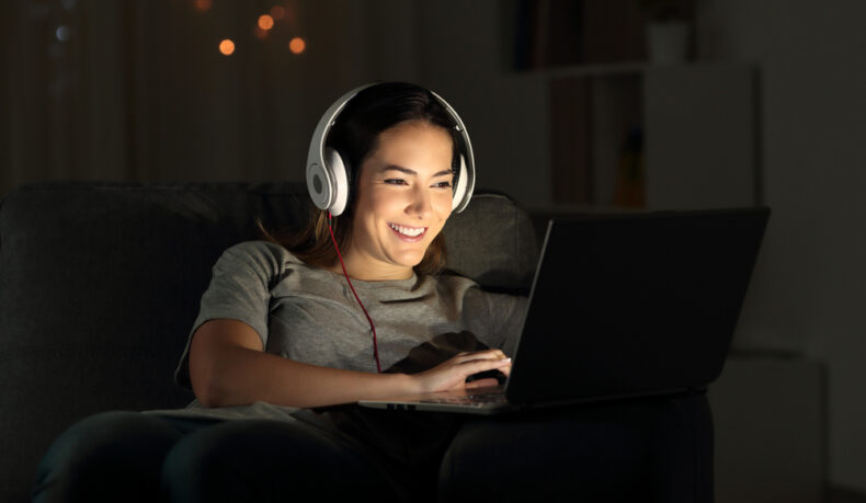 O femeie tânără, cu o pereche de căști pe cap, zâmbește și se uită la ecranul unui laptop noaptea, într-o cameră