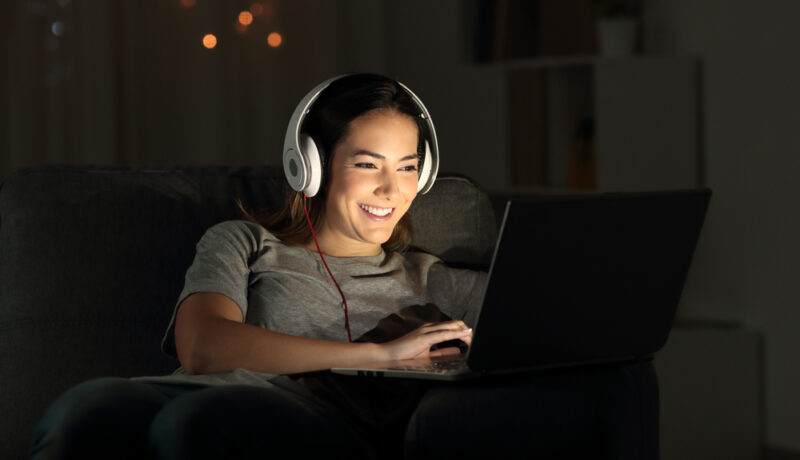 O femeie tânără, cu o pereche de căști pe cap, zâmbește și se uită la ecranul unui laptop noaptea, într-o cameră