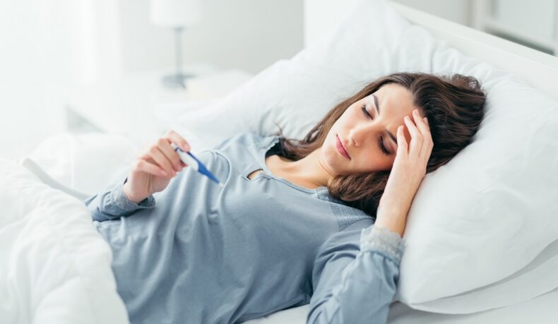 Femeie care stă în pat și se uită la termometru, febra fiind unuldintre cele mai coune simptome ale infecției cu citomegalovirus