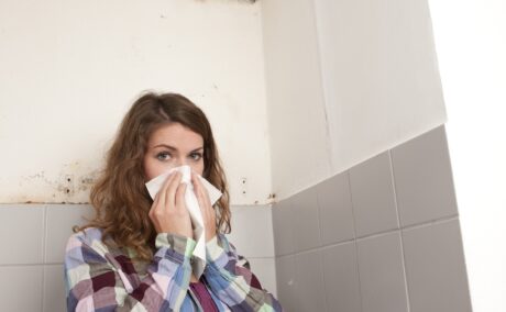 Femeie cu șervețel la nas și mucegai pe peretele din baie, în fundal, sugestiv pentru alergia la mucegai