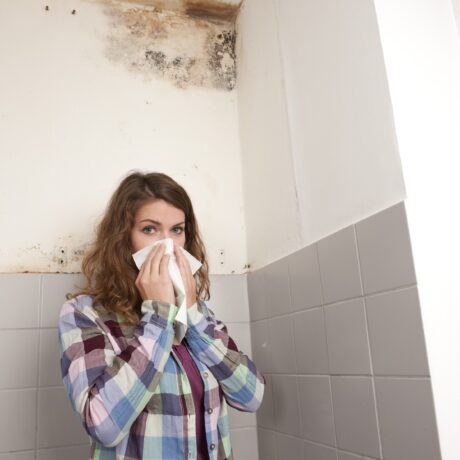 Femeie cu șervețel la nas și mucegai pe peretele din baie, în fundal, sugestiv pentru alergia la mucegai