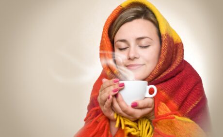 Femeie înfășurată într-o pătură care bea o ceașcă de cafea, sugestiv pentru cât de indicat e să bei cafea când ești răcit