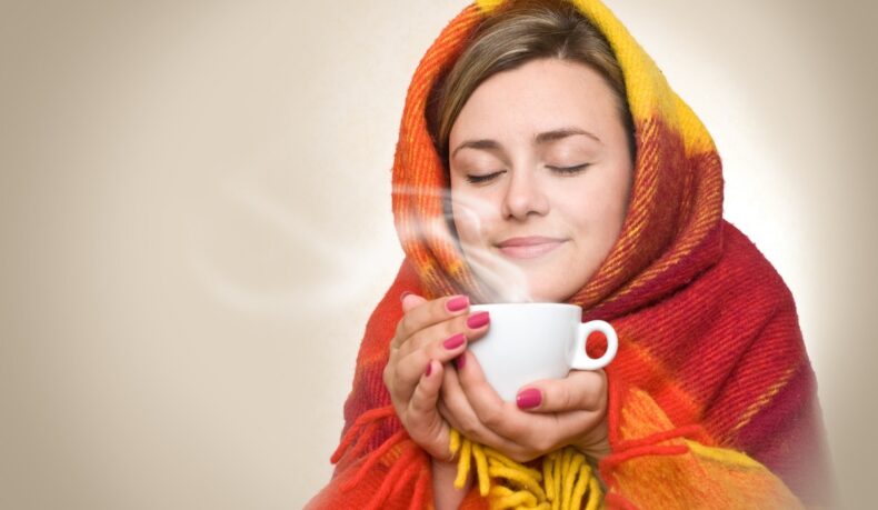 Femeie înfășurată într-o pătură care bea o ceașcă de cafea, sugestiv pentru cât de indicat e să bei cafea când ești răcit