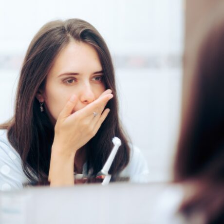 O femeie tânără ține mână la gură din cauza unei dureri gingivale, în timp ce privește în oglindă