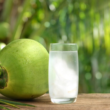 Un pahar cu apă de cocos, lângă o nucă de cocos cu coajă verde, pe o masă din lemn