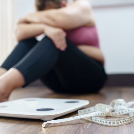 O femeie tânără supărată stă pe podea lângă un cântar și un centimetru de croitorie