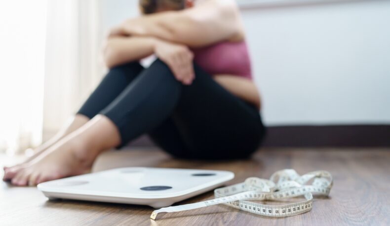 O femeie tânără supărată stă pe podea lângă un cântar și un centimetru de croitorie