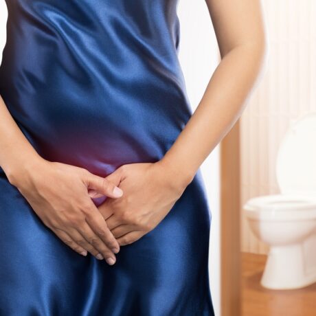 Femeie cu mâinile în zona intimă și toaletă în fundal, sugestiv pentru infecții urinare