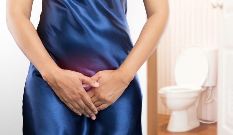 Femeie cu mâinile în zona intimă și toaletă în fundal, sugestiv pentru infecții urinare