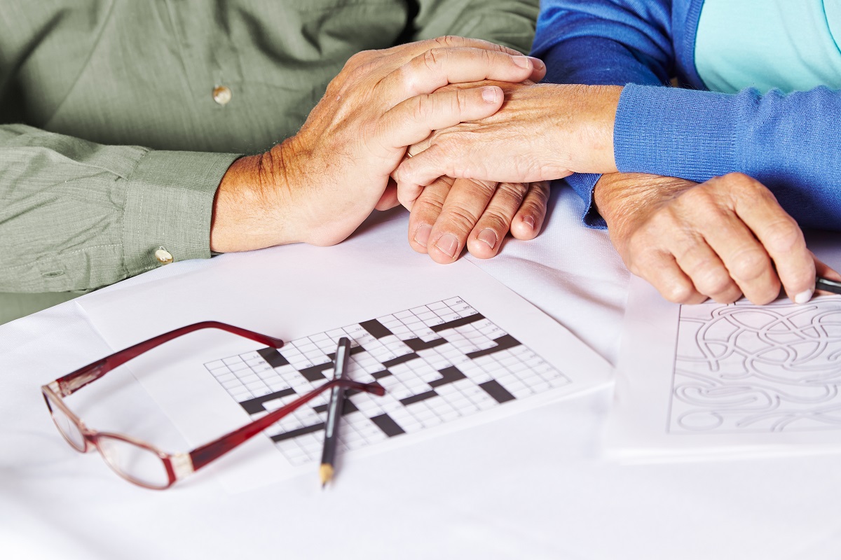 Vârstnici care se țin de mâini și un rebus nerezolvat pe masă, sugestiv pentru Alzheimer sau alte tipuri de demență