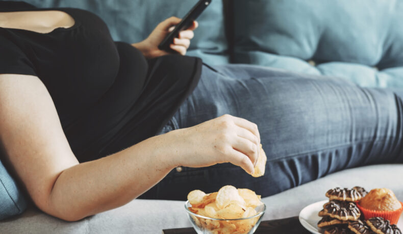 O femeie cu obezitate, întinsă pe canapea, mănâncă din două vase chipsuri și brioșe