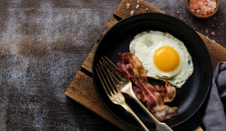 O tigaie cu un ou, bacon și două furculițe, pusă pe un suport din lemn, lângă un vas cu sare