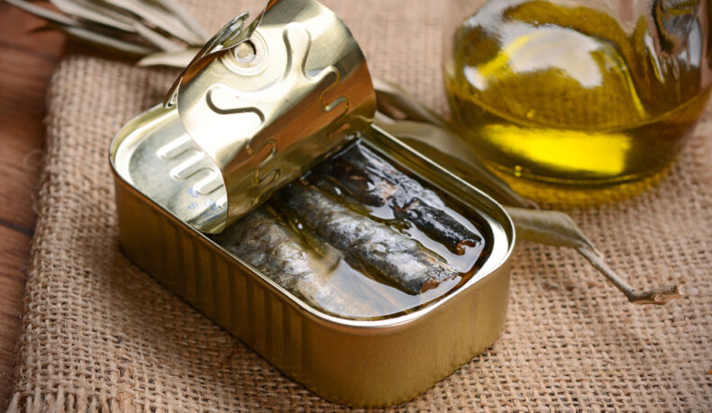 O conservă desfăcută, cu sardine, lângă o cană cu ulei