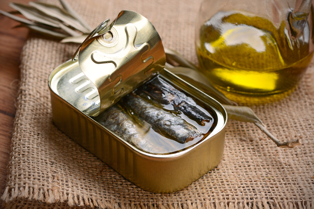 O conservă desfăcută, cu sardine, lângă o cană cu ulei