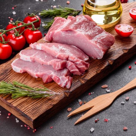 Carne de porc pe un tocător, cu roii și condimente în jur, sugestiv pentru câte calorii are carnea de porc