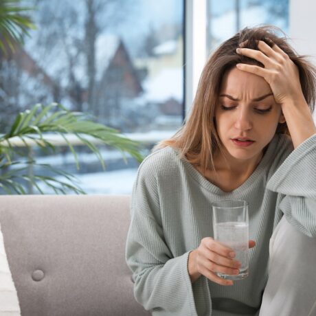 Femeie cu durere de cap care bea un pahar cu apă și medicamente, sugestiv pentru ce să faci dacă ai băut prea mult