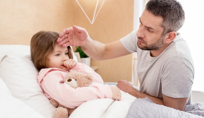 Fetiță bolnavă care stă în pat și tușește, iar tatăl îi verifică temperatura cu mâna pe frunte. Copiii sunt o categorie expusă la complicațiile gripei