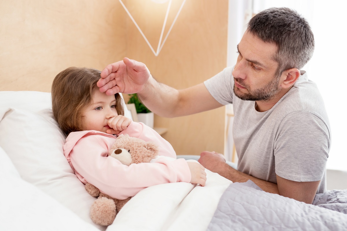 Fetiță bolnavă care stă în pat și tușește, iar tatăl îi verifică temperatura cu mâna pe frunte. Copiii sunt o categorie expusă la complicațiile gripei