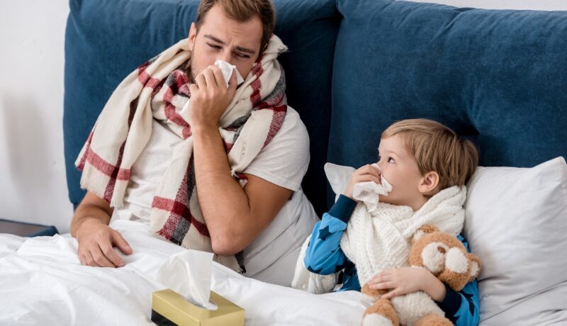 Tată și fiu în pat, cu gripă sau răceală, își suflă nasul
