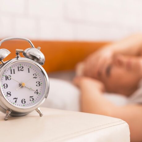 Ceas pe noptieră și femeie în fundal care nu poate să doarmă, sugestiv pentru motivele care explică de ce dormim mai puțin când îmbătrânim