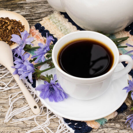 O ceașcă cu cafea de cicoare, lângă: flori de cicoare, un vas, o farfurie din lemn cu rădăcini de cicoare și o lingură din lemn