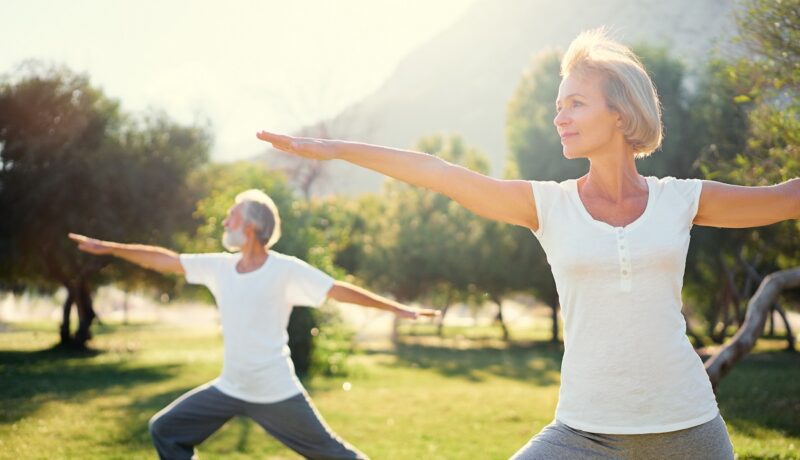 Cuplu de vârstnici care face yoga în parc, sugestiv pentru un stil de viață sănătos