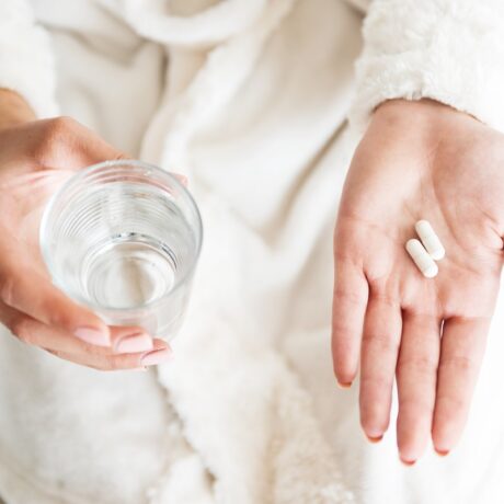 Femeie cu două capsule albe în palmă și un pahar cu apă în cealaltă mână, sugestiv pentru colostrul sub formă de capsule