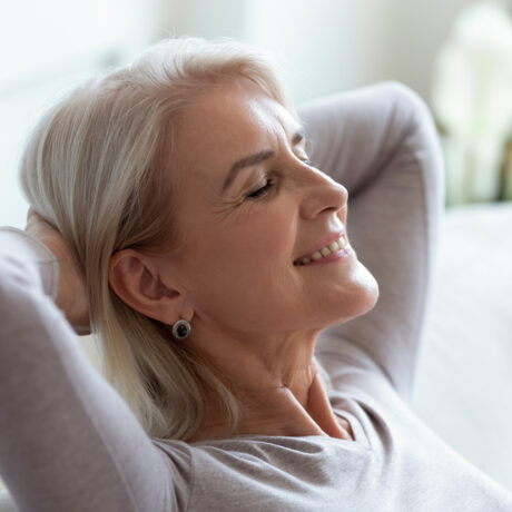 O femeie în vârstă zâmbește și își ține mâinile sub cap, pe o canapea