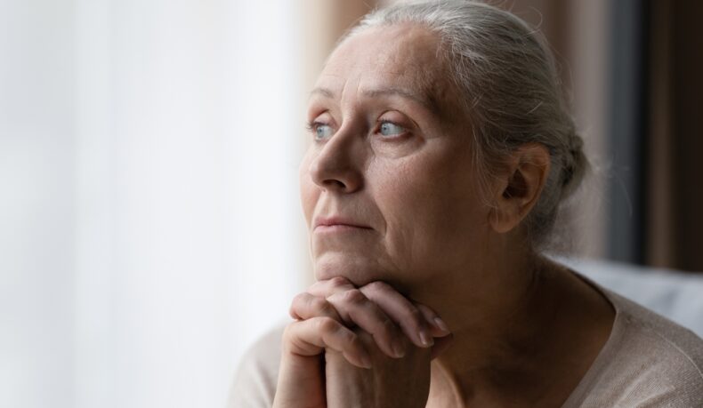 O femeie în vârstă privește îngrijorată pe fereastră