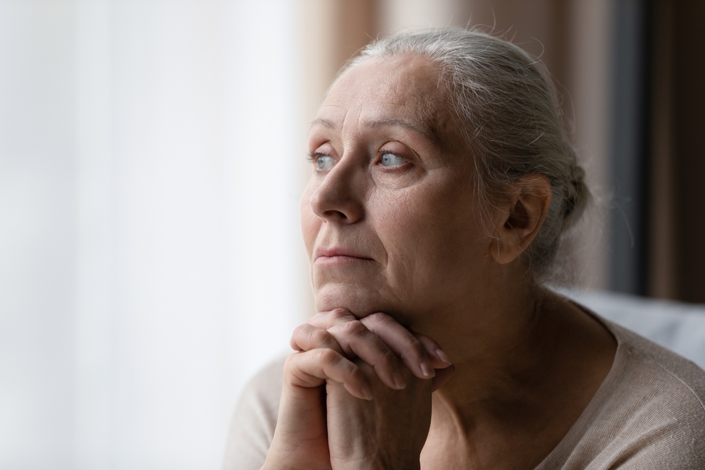 O femeie în vârstă privește îngrijorată pe fereastră
