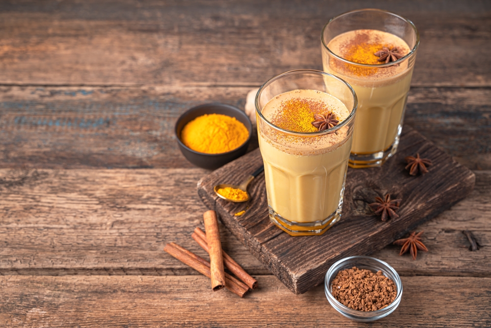 Două pahare cu ceai latte de turmeric, lângă: anason, batoane de scorțișoară și un vas cu pudră de turmeric, puse pe un suport din lemn