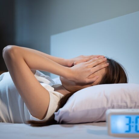O femeie stă întinsă în pat cu mâinile pe față, lângă un ceas digital