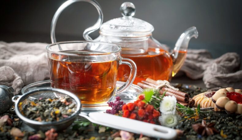 O ceașcă și un ceainic cu ceai, între plante medicinale, fursecuri și scorțișoară